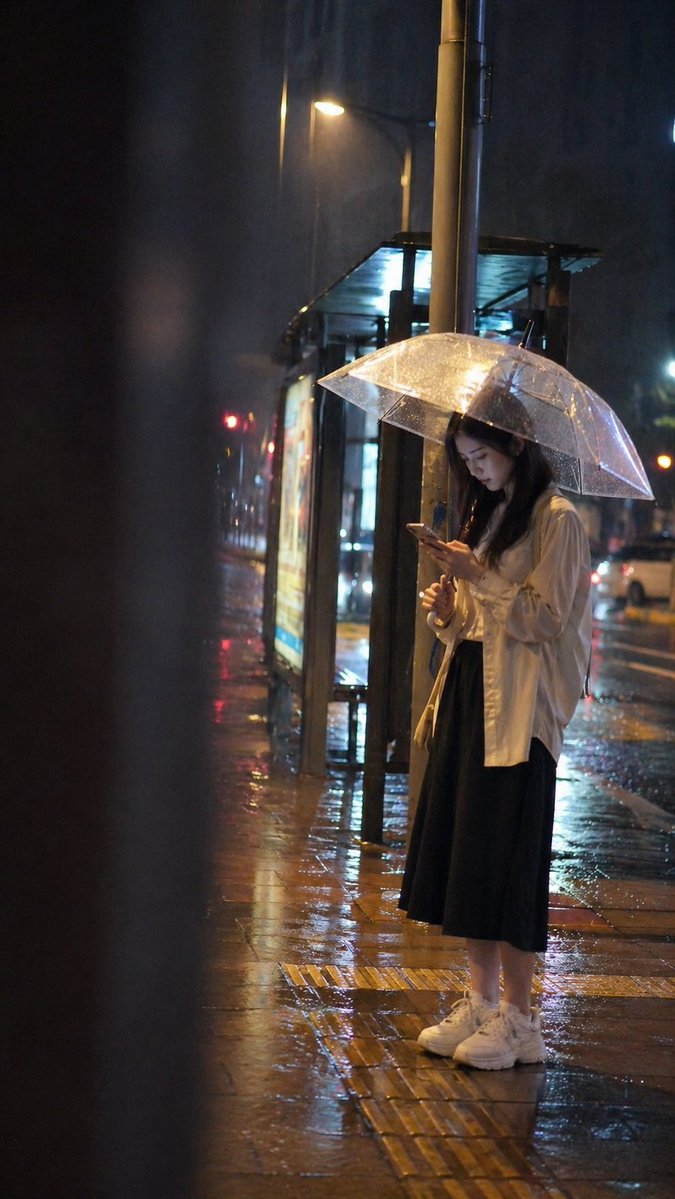 At a city bus stop on a rainy night, a young adult woman stands alone…